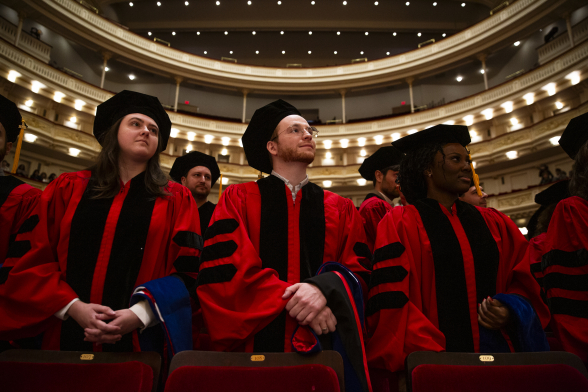 Students in graduation regalia stand in an auditorium