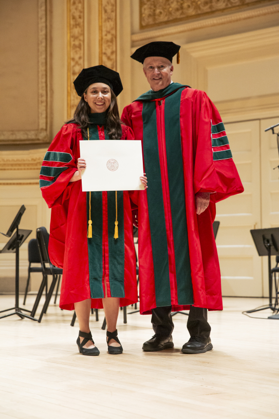 A graduate stands with a school dean on an ornate stage