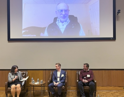 A panel consists of one woman, two younger men sitting on stage and an older man on screen.