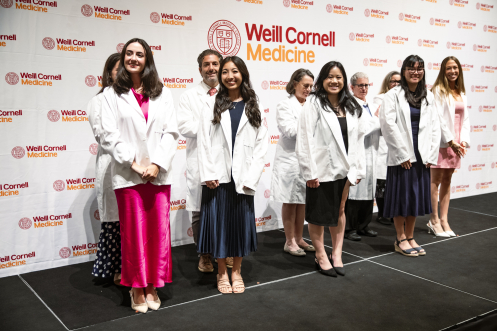 Four students wearing white coats standing in front of four faculty members