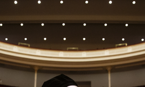 Students wearing graduation regalia stand in an ornate auditorium