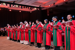 students wearing commencement regalia reading and raising their right hands