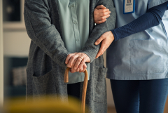 Elder woman using walking cane at nursing home with nurse holding hand for support.