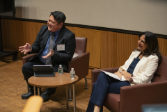 An East Asian man wearing a dark gray suit and tie and a South Asian woman with long black hair and wearing a white suit. Both smiling at the audience. 