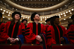 Students wearing graduation regalia stand in an ornate auditorium